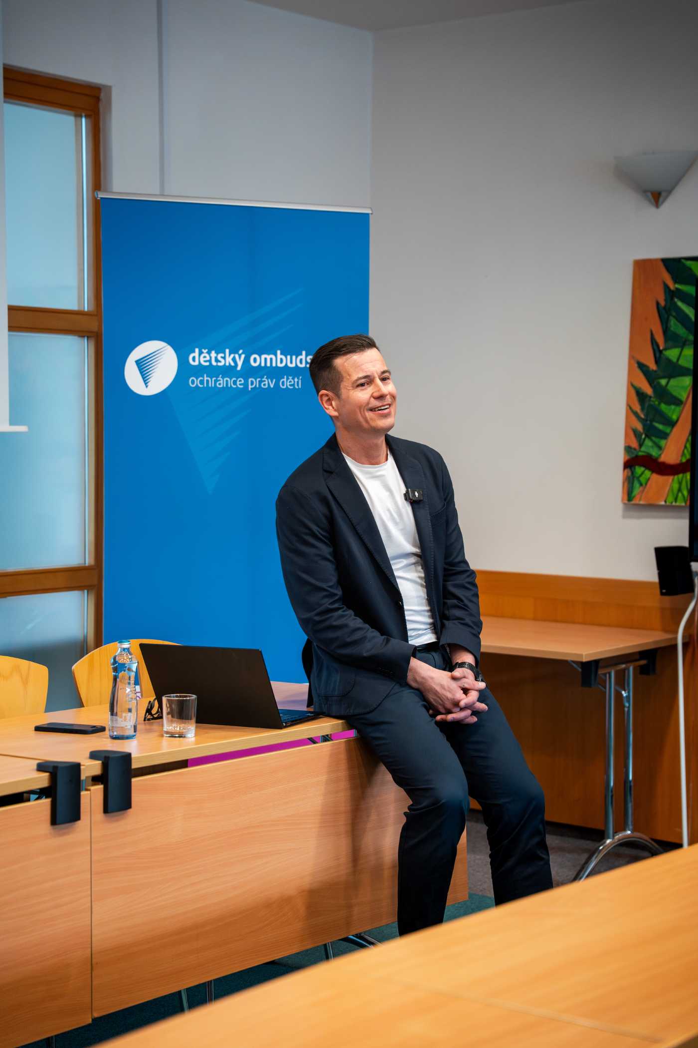 Dětský ombudsman Martin Beneš sedí na stole před bannerem s logem dětského ombudsmana a mluví k novinářům na briefingu.