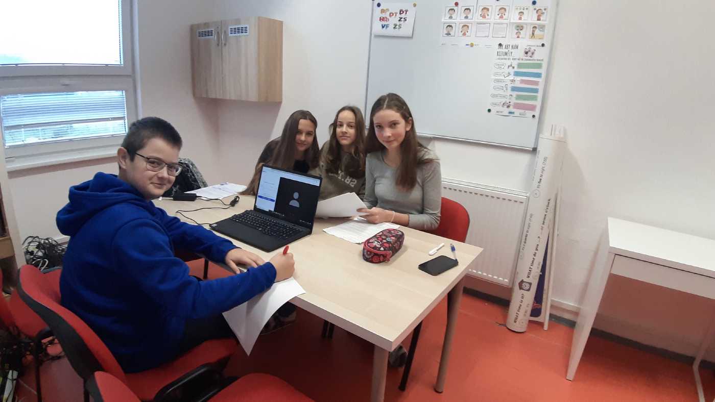 U stolu s počítačem sedí při online setkání tři dívky a jeden chlapec.