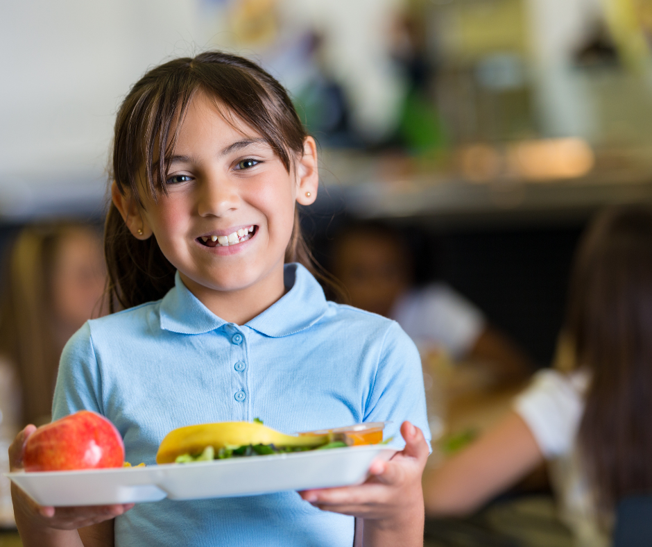 Fotka zachycuje dívku ve školní jídelně. V ruce drží tác s jídlem. 