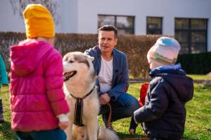Dětský ombudsman Martin Beneš dřepí, přidržuje svého huskyho a v popředí jsou dvě malé děti, které stojí čelem k němu.